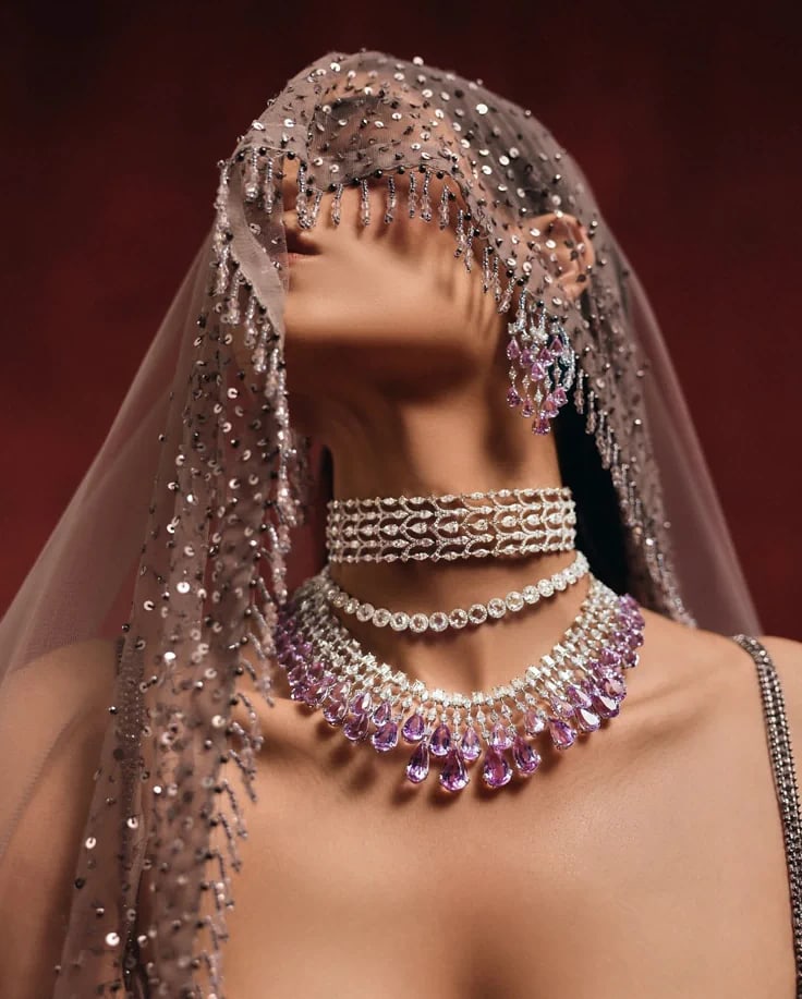 A woman in a beaded veil against a deep red ground, wearing a diamond choker and a necklace of pink teardrop stones.